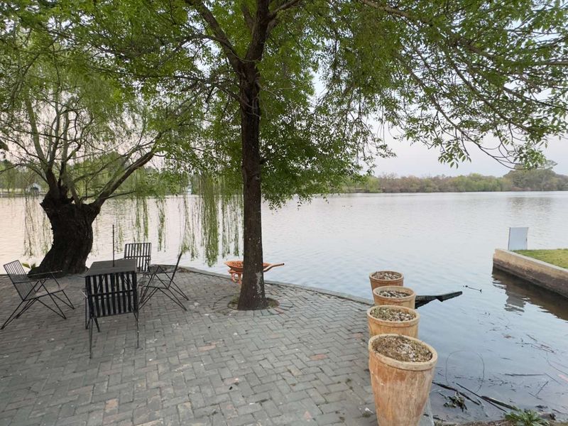 A serene waterfront patio area featuring paved brick flooring, mature trees, and direct access to the lake, offering a peaceful outdoor retreat.
