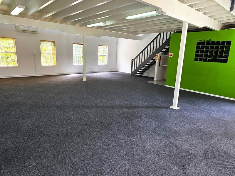 Spacious open-plan office area featuring carpeted flooring, exposed ceiling beams, and a staircase leading to an upper level.