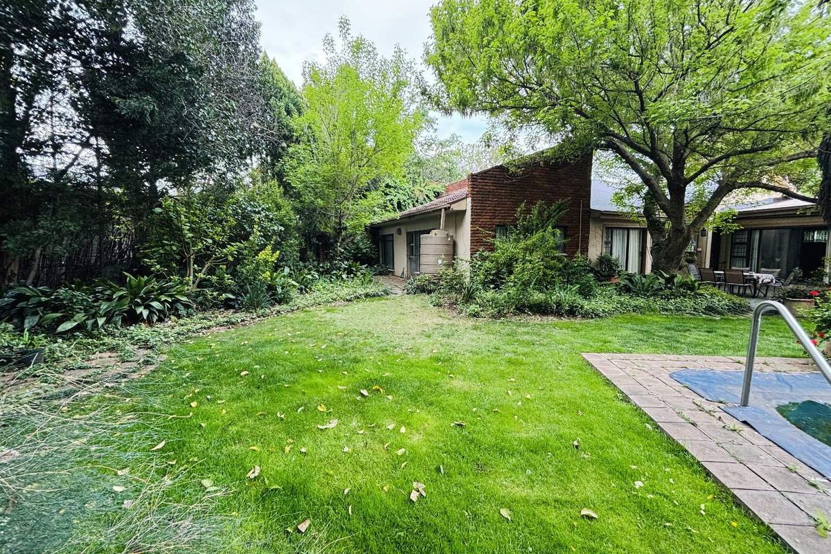 A spacious rear garden featuring a lush lawn, mature trees, a swimming pool with paved surround, and a brick-clad exterior wall with sliding door access.