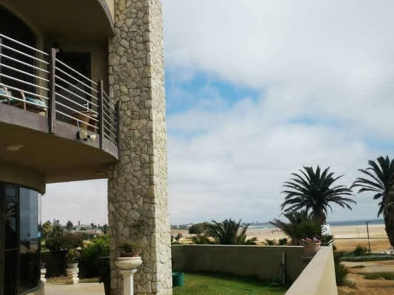 Rear exterior view featuring a stone-clad chimney, a balcony with metal railings, a tiled patio area, and a lawn overlooking the coastal landscape.