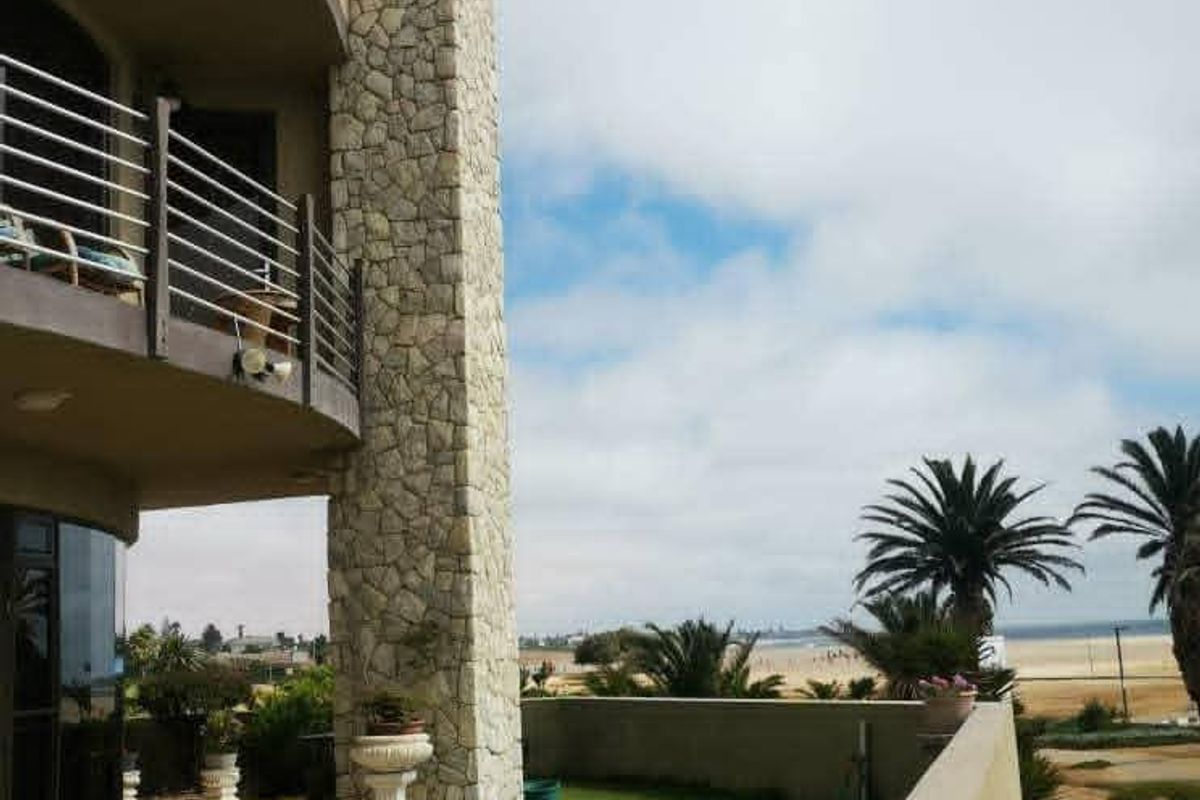 Rear exterior view featuring a stone-clad chimney, a balcony with metal railings, a tiled patio area, and a lawn overlooking the coastal landscape.