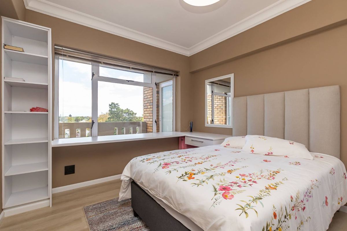 A well-lit bedroom featuring a built-in shelving unit, a window-side desk, and a large window with views, finished with neutral wall tones.