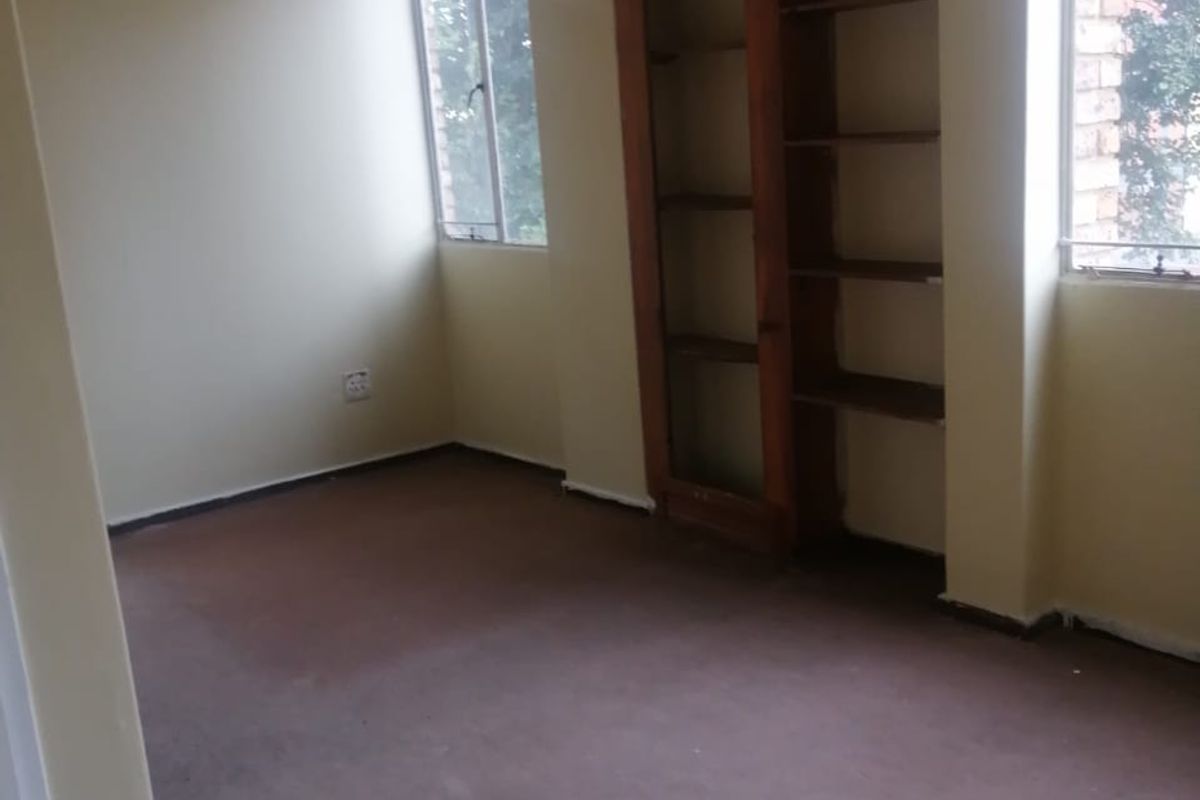 An empty bedroom featuring built-in wooden shelving, carpeted flooring, and large windows providing natural light. The space is ready for renovation or staging.