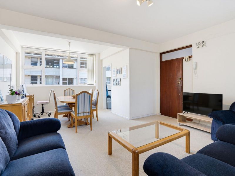 An open-plan living and dining area featuring neutral carpet flooring, large windows for natural light, and a secure timber entrance door.