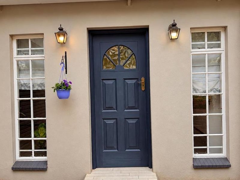 A well-maintained front entrance featuring a dark blue panelled door with leaded glass, flanked by two sash windows and exterior wall lighting.