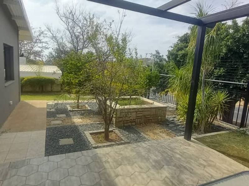 A covered patio area featuring modern paving, a stone planter box, and a pergola, overlooking a landscaped garden with security fencing.