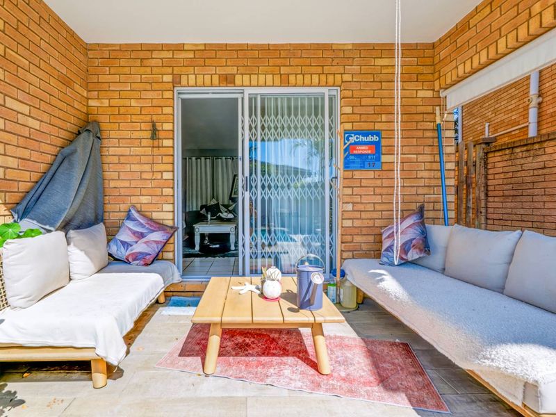 A covered patio area featuring exposed brick walls, tiled flooring, and a sliding glass door with a security gate providing access to the interior.