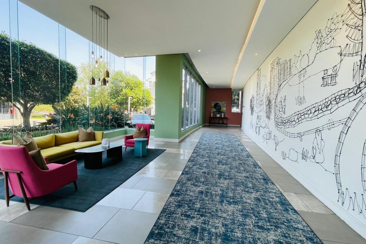 A modern, bright apartment lobby or lounge area featuring floor-to-ceiling glass windows, tiled flooring, and a large decorative wall mural.