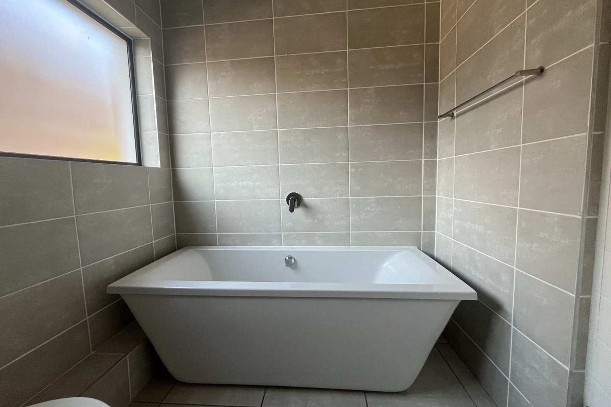 A modern bathroom featuring a clean freestanding bathtub, wall-mounted faucet, and neutral-toned wall and floor tiling with a frosted window for privacy.