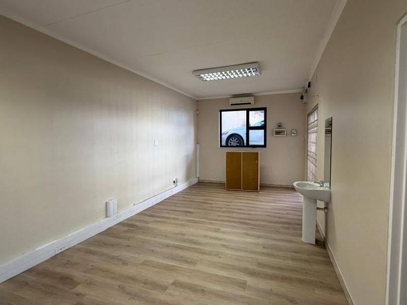 A clean, empty office space featuring laminate flooring, a wall-mounted air conditioning unit, and a convenient pedestal sink.