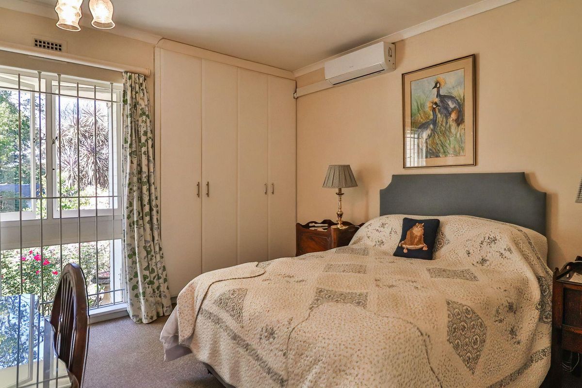 A bedroom featuring built-in wardrobes, a wall-mounted air conditioning unit, and a large window with security bars overlooking the garden.