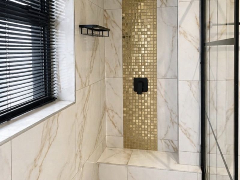 Modern walk-in shower featuring marble-look wall tiles, a gold mosaic feature strip, built-in bench, and contemporary black fixtures.