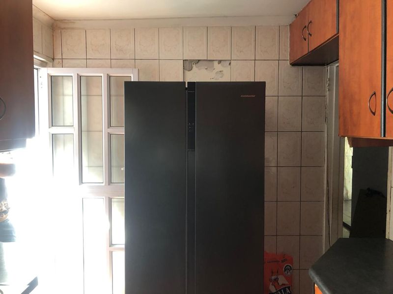 A kitchen area featuring wooden cabinetry and tiled splashbacks, currently showing visible wall damage behind the refrigerator space.