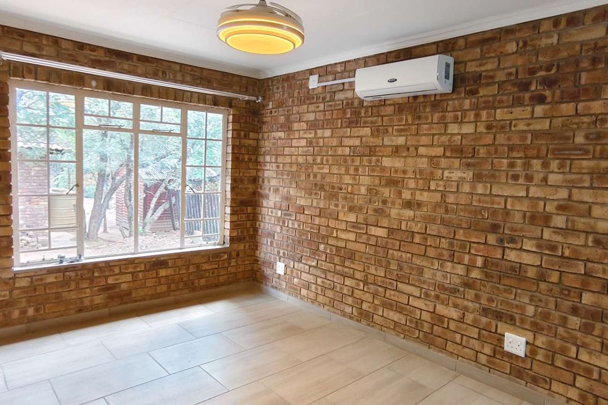 A bright bedroom featuring exposed brick walls, tiled flooring, a large window, and a wall-mounted air conditioning unit.