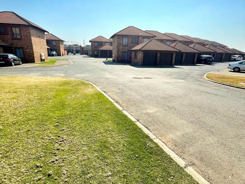 A view of the cluster complex entrance featuring facebrick facades, tiled roofs, paved driveways, and multiple garage units within a well-maintained estate setting.