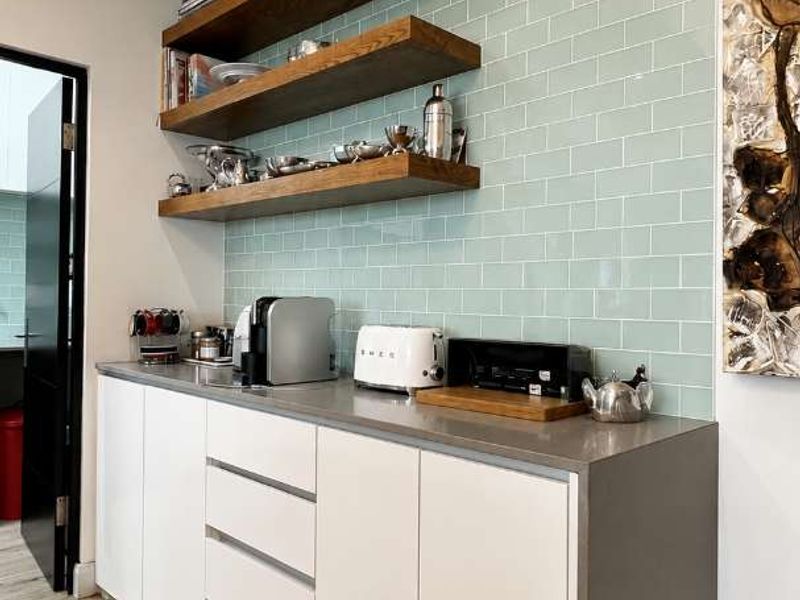 Modern kitchen area featuring sleek white cabinetry, stone countertops, a light green subway tile backsplash, and stylish floating timber shelving.