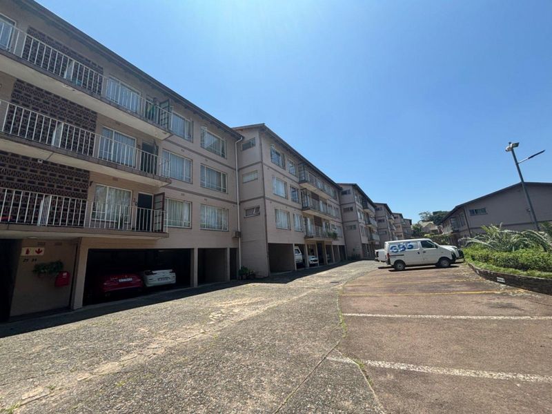 Exterior view of a multi-story apartment complex featuring covered parking bays, balconies, and a paved driveway area.