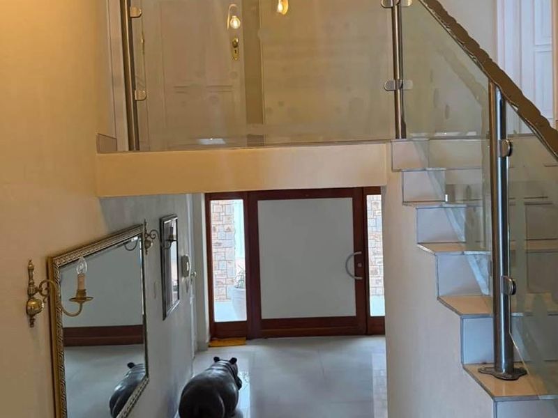 A bright entrance hall featuring a modern staircase with a glass balustrade, stainless steel handrail, and polished tiled flooring leading to the front door.
