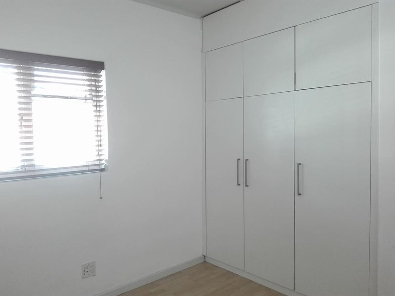 A clean, empty bedroom featuring white built-in cupboards, light wood-look flooring, and a window fitted with venetian blinds.
