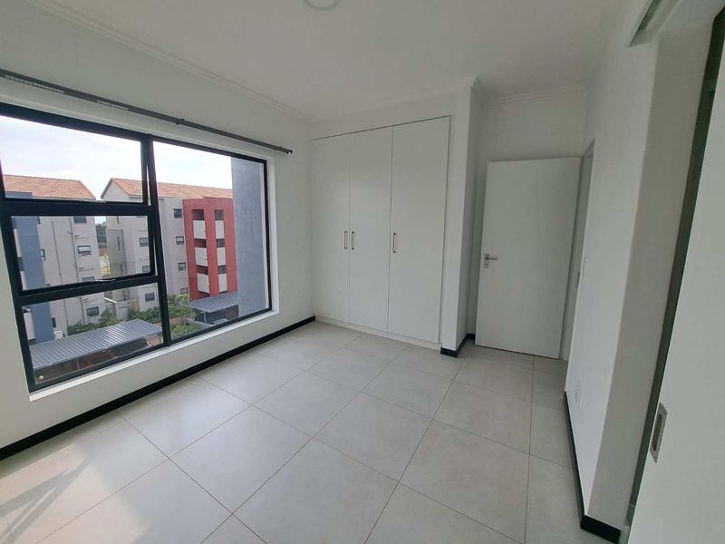 A bright, empty bedroom featuring neutral tiled flooring, built-in cupboards, and a large window providing natural light and views of the complex.