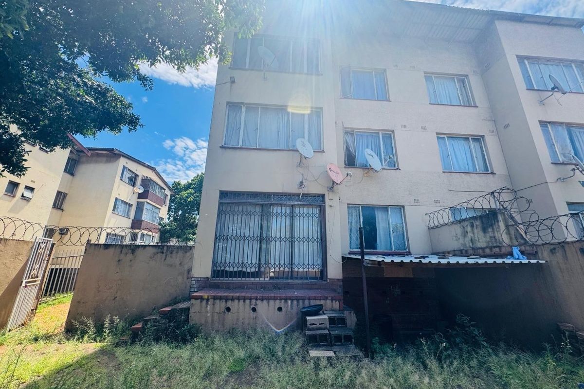 Rear view of a multi-story apartment building featuring security gates, razor wire fencing, and an overgrown garden area requiring maintenance.