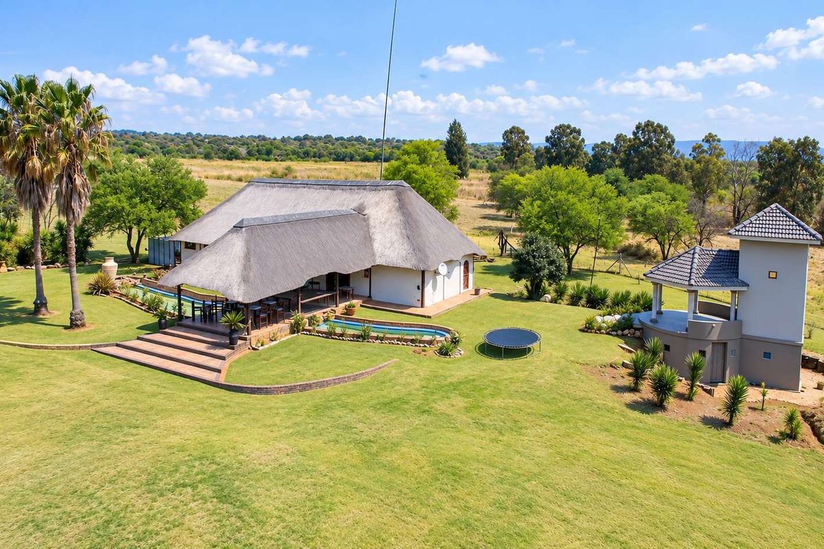 Aerial view of a lodge property featuring a thatched roof main building, swimming pool, patio area, and a secondary structure set in a large garden.