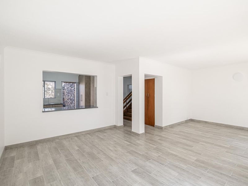 A bright, empty living area featuring modern grey tiled flooring, a convenient pass-through window to the kitchen, and access to a staircase.