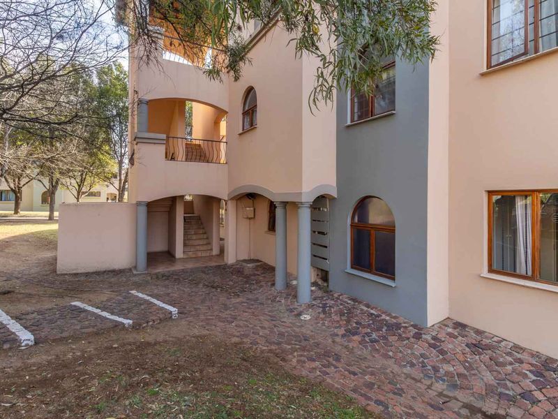 Exterior view of an apartment building featuring arched architectural details, a covered balcony, staircase, and cobblestone paving in a landscaped setting.