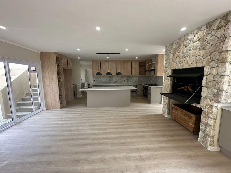 Spacious open-plan living and kitchen area featuring modern wood-finish cabinetry, a central island, and a striking stone-clad fireplace with a built-in braai.