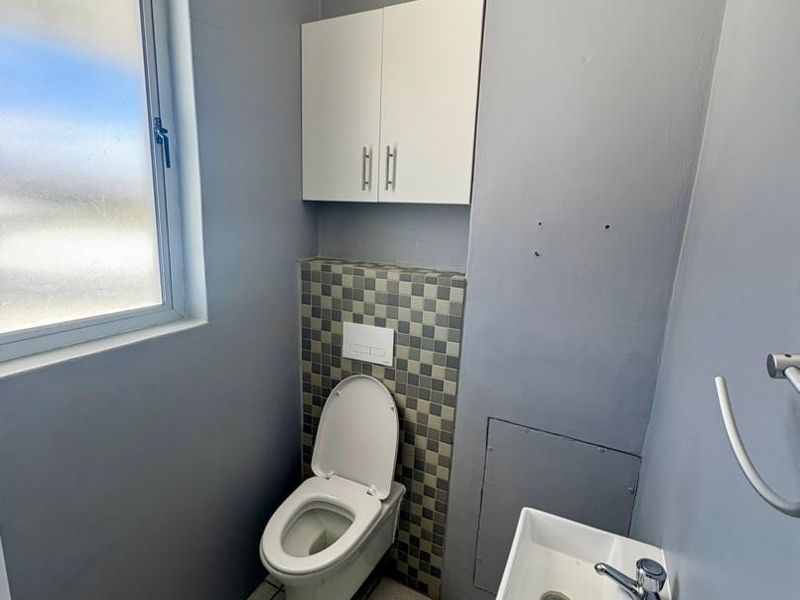 A compact office restroom featuring a wall-mounted toilet with concealed cistern, a small wall-mounted basin, and neutral grey wall finishes.