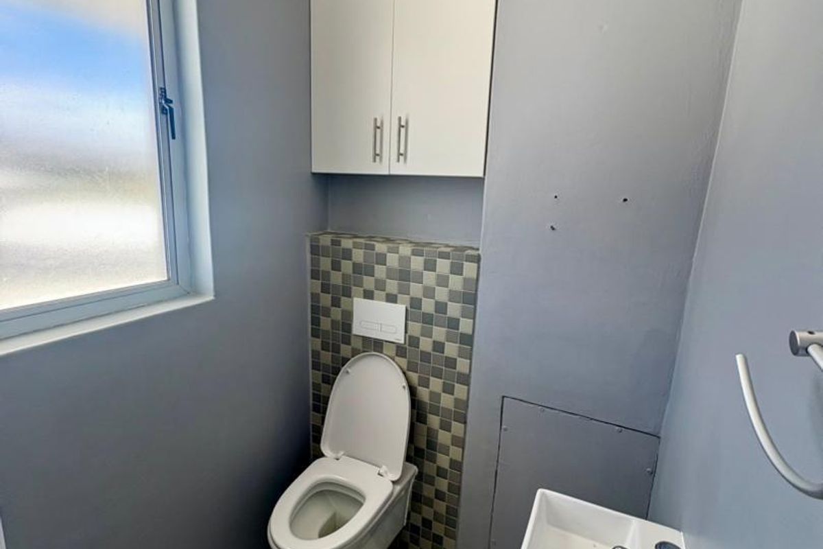 A compact office restroom featuring a wall-mounted toilet with concealed cistern, a small wall-mounted basin, and neutral grey wall finishes.