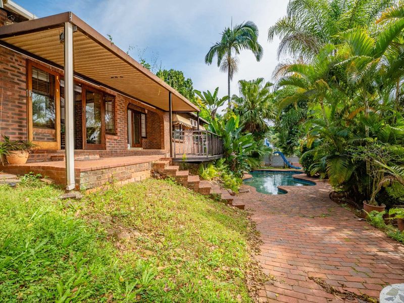 Rear exterior view featuring a brick facade, covered patio area, swimming pool, and established garden with brick pathways.
