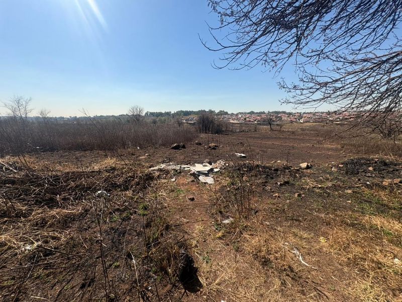 An undeveloped residential land plot featuring dry vegetation and scattered debris, offering an open view of the surrounding neighborhood.