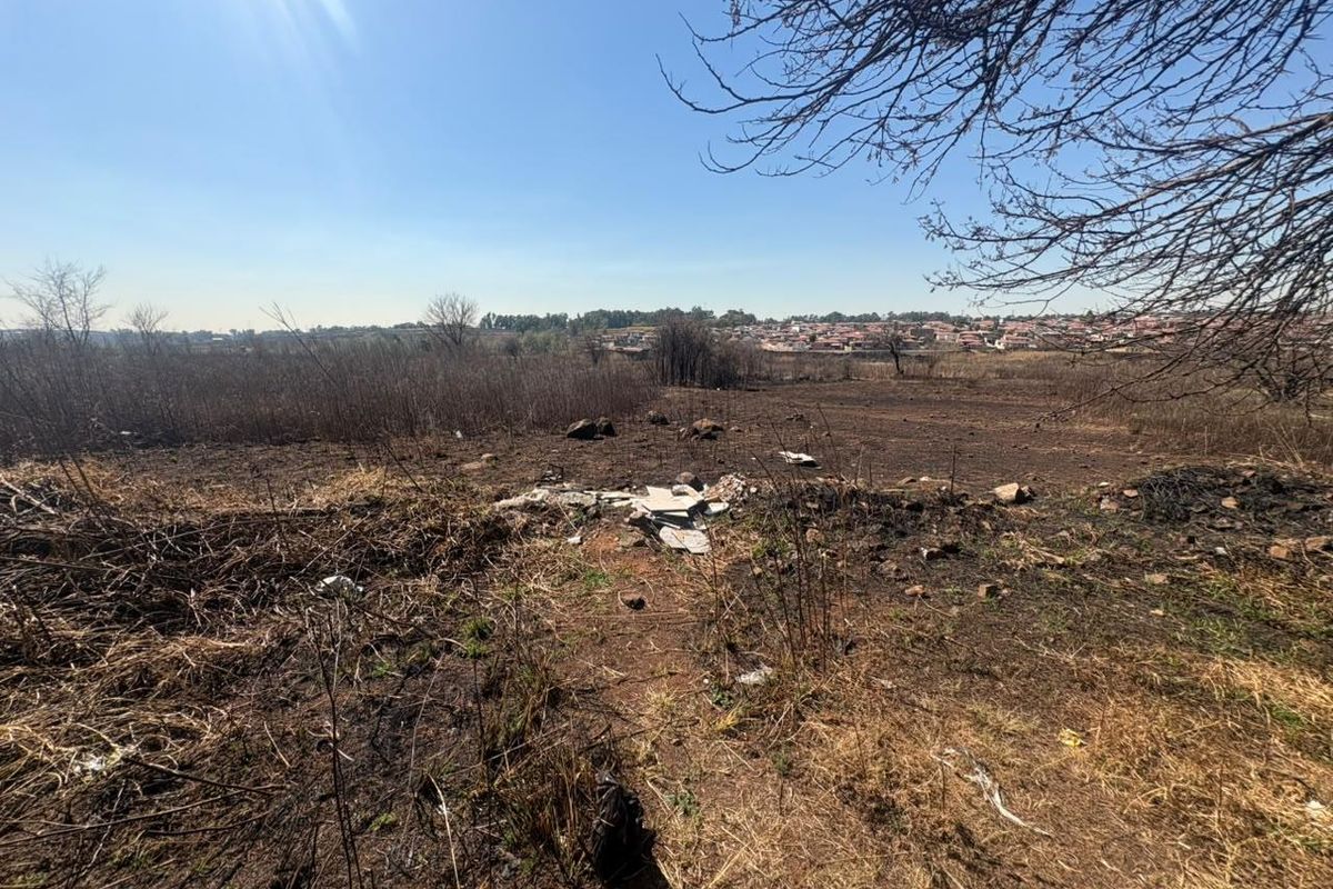 An undeveloped residential land plot featuring dry vegetation and scattered debris, offering an open view of the surrounding neighborhood.