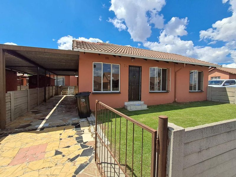 Front view of a single-story townhouse featuring a tiled roof, attached carport, paved driveway, and a small front garden with boundary walling.