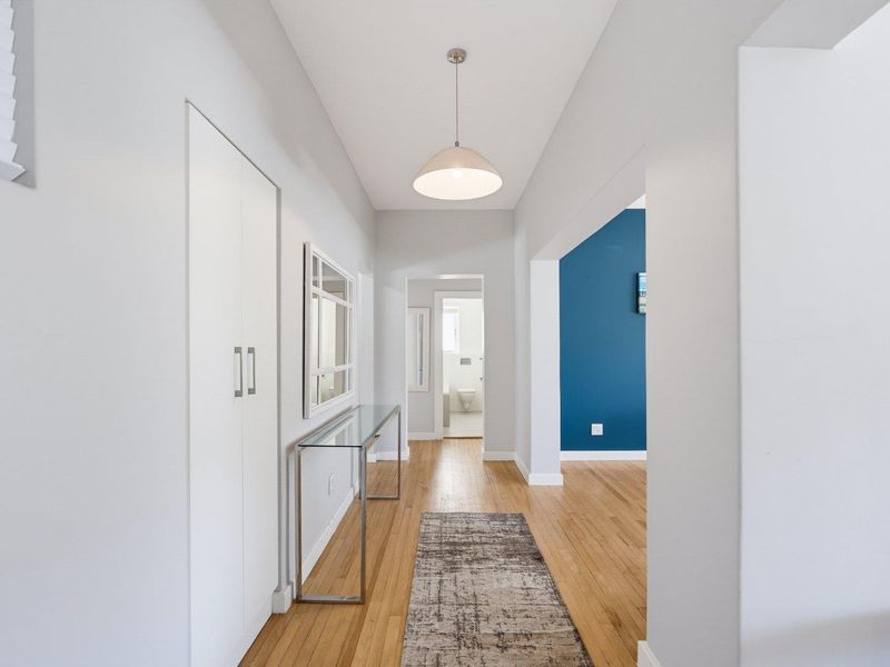 A bright, well-maintained hallway featuring hardwood flooring, a modern pendant light, and a built-in storage cupboard, leading towards a bathroom and living area.