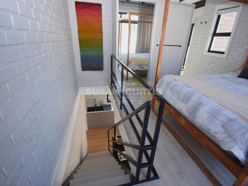 A loft-style bedroom featuring painted brick walls, laminate flooring, and a metal staircase overlooking a small lower-level kitchenette area.