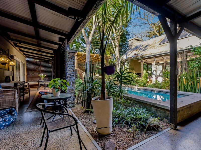 A covered patio area overlooking a private swimming pool and landscaped garden, featuring stone accents and exposed timber beams.