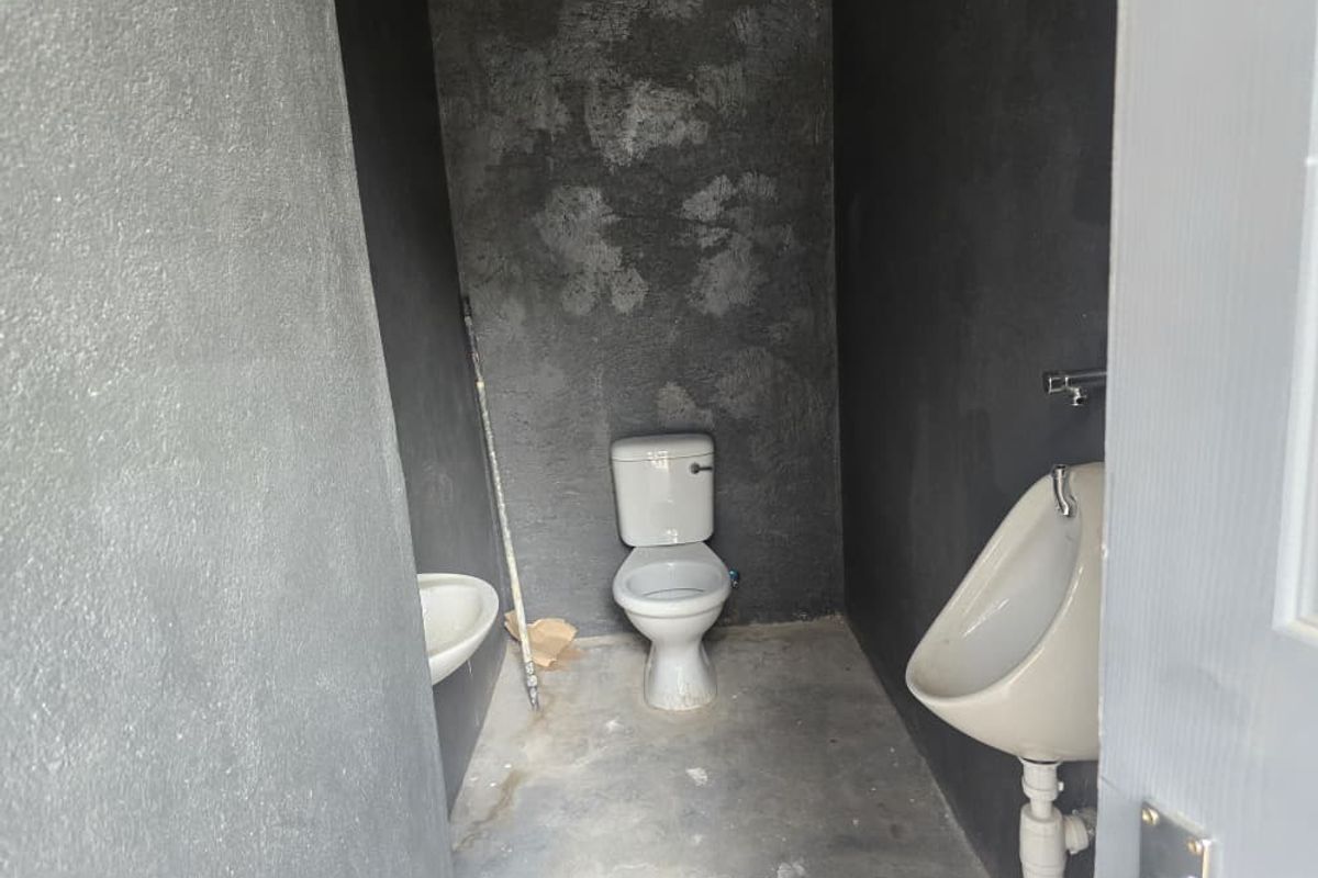 A functional restroom facility featuring a toilet, wall-mounted urinal, and basin, finished with raw, dark-textured walls and concrete flooring.