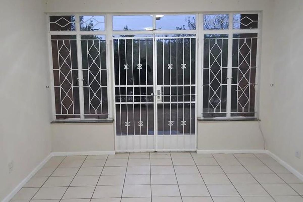 An empty living room featuring neutral tiled flooring, a central ceiling light, and secure metal security gates and window bars.