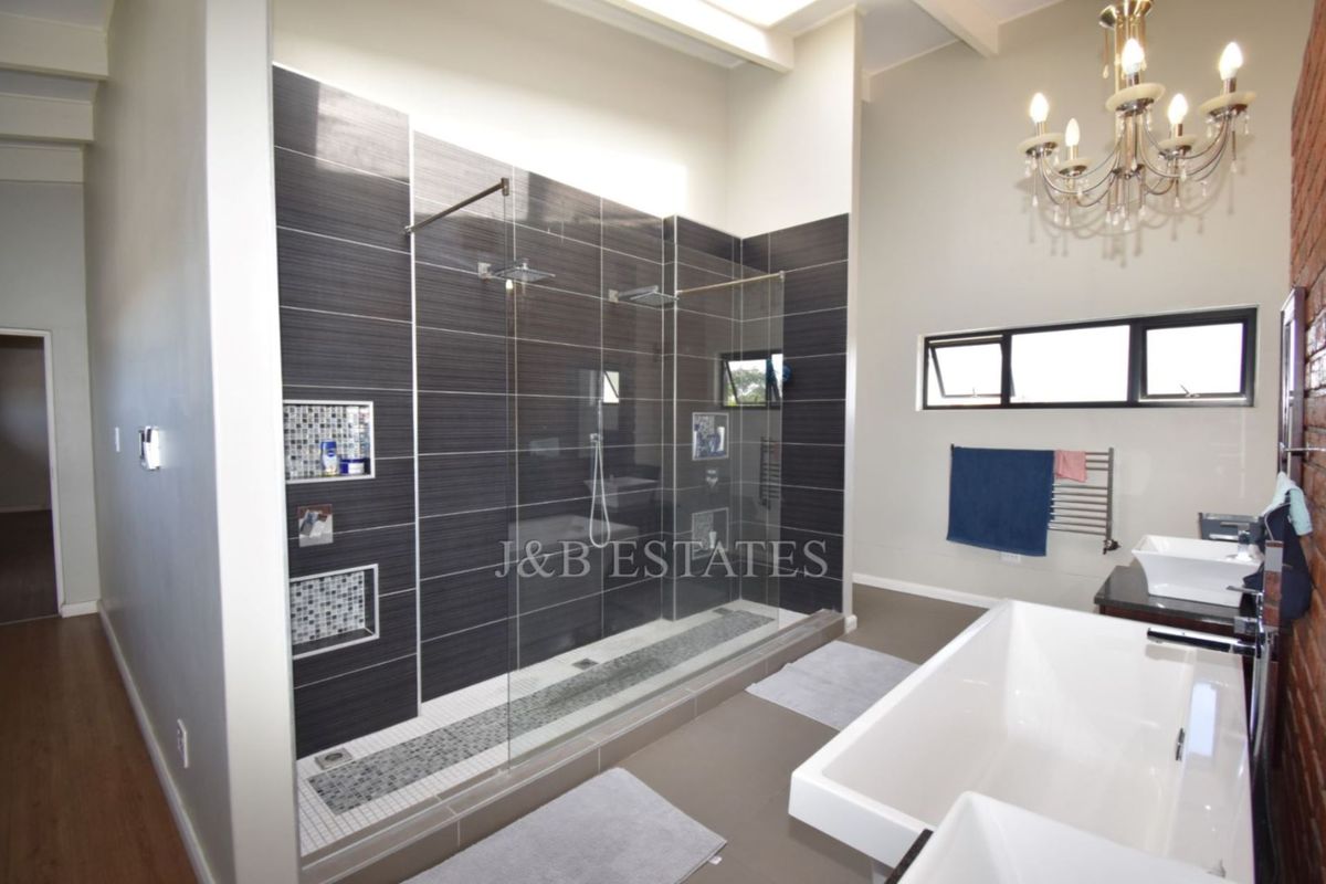 Modern bathroom featuring a large walk-in shower with dark tiling, a freestanding bathtub, and a double vanity, complemented by high ceilings and exposed brick.