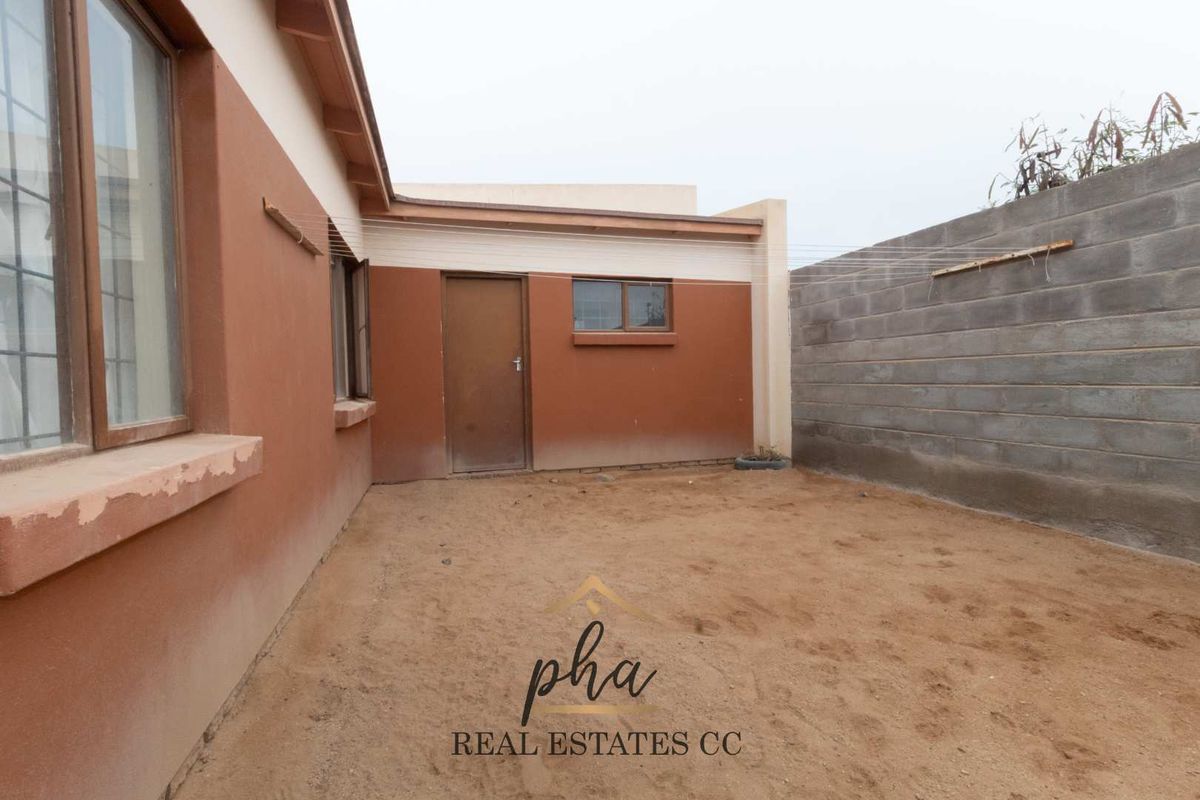 Rear exterior view of the property featuring a sandy yard, exposed concrete block boundary wall, and painted exterior facade with window and door access.