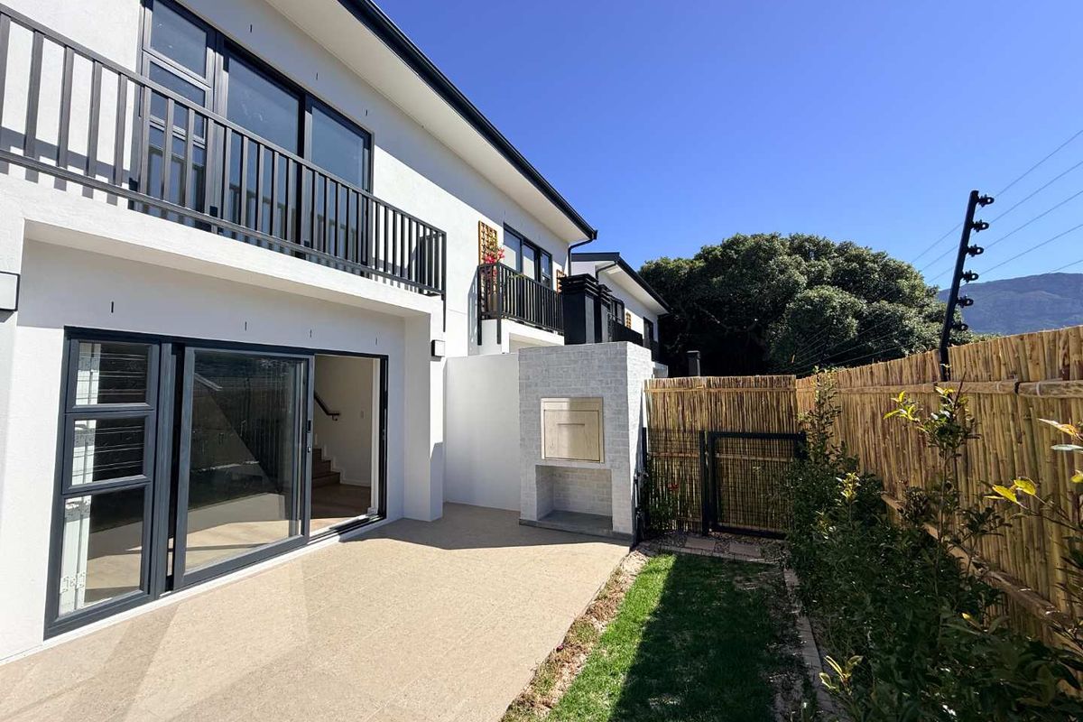 Rear exterior view of a modern townhouse featuring a paved patio, built-in braai, balcony with metal railings, and secure perimeter fencing.