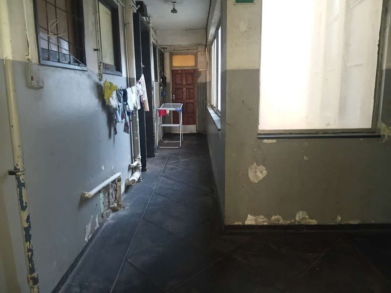A narrow interior hallway featuring tiled flooring, large windows, and exposed piping, currently showing significant wear to the wall finishes.
