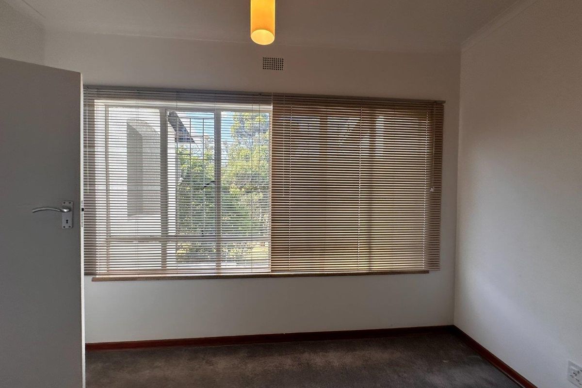 A bright, empty bedroom featuring neutral walls, carpeted flooring, and large windows fitted with venetian blinds.