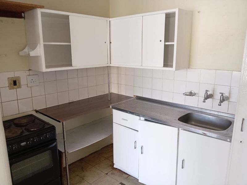 A compact kitchen featuring white cabinetry, a stainless steel sink, tiled splashback, and a freestanding stove, requiring cosmetic updates and repairs.