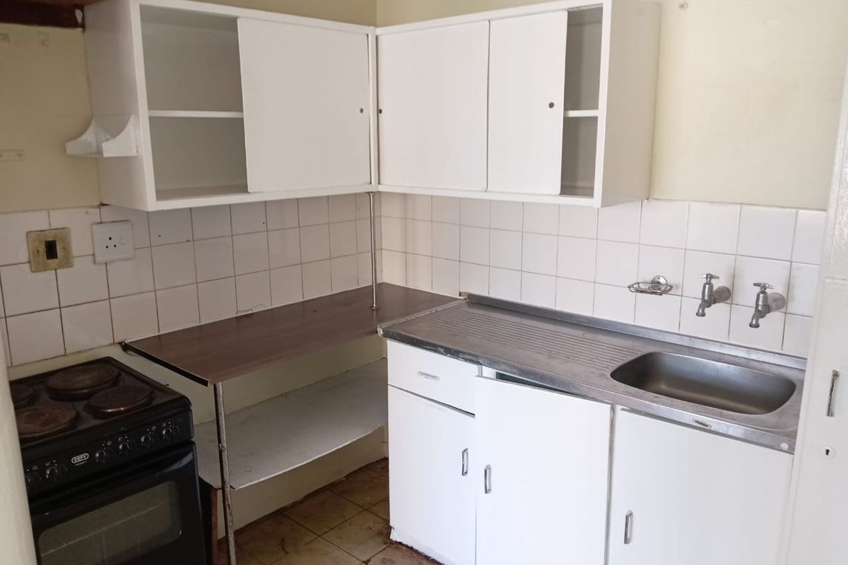 A compact kitchen featuring white cabinetry, a stainless steel sink, tiled splashback, and a freestanding stove, requiring cosmetic updates and repairs.