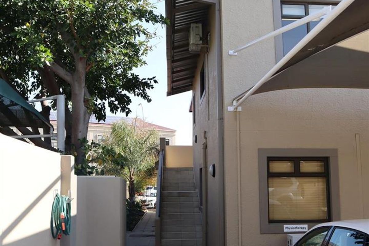 Exterior view of the office building side profile, featuring paved walkway, external staircase, and shaded parking area.