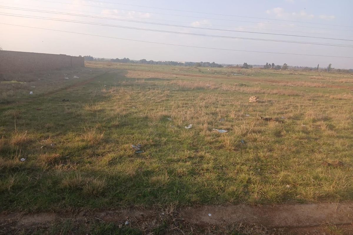 A view of a vacant residential plot featuring open grass land, a boundary wall, and overhead power lines.