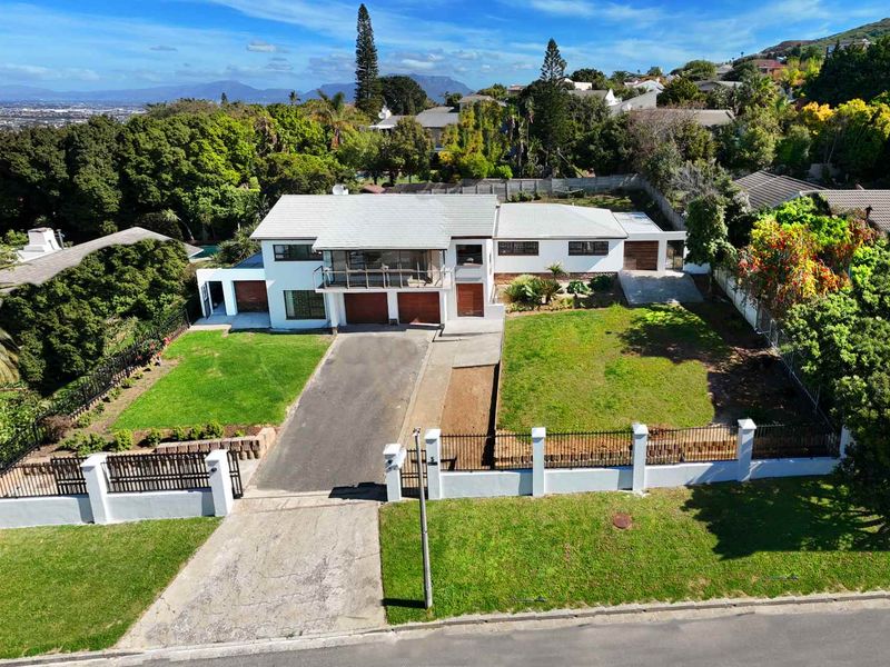 Aerial view of a residential property featuring a double garage, balcony, paved driveway, and a spacious front garden enclosed by a perimeter wall.